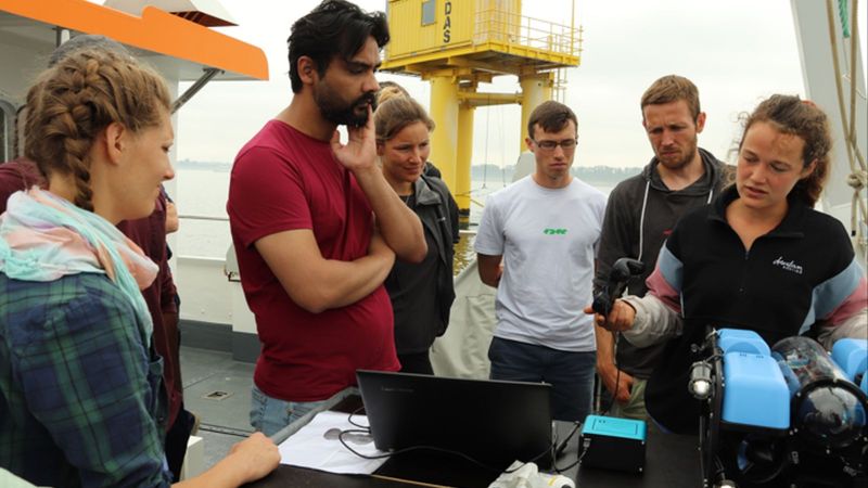 Das Bild zeigt die Oberkörprer von sechs jungen Menschen, die an Bord eines Schiffes sind und sich technische Ausrüstung ansehen.