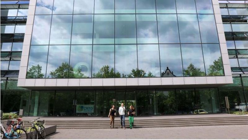 Hausfassade mit viel Glas davor drei Menschen und ein Fahrradständer.